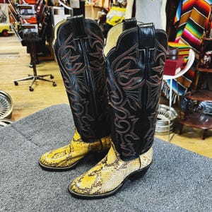 Könnte beinhalten: Ein Paar Cowboy-Stiefel mit schwarzen Lederoberteilen und aufwendigen Nähten in einem rötlich-braunen Farbton. Der untere Teil der Stiefel weist ein gelbes und beiges Schlangenmuster auf. Die Stiefel werden auf einer grauen Oberfläche präsentiert.