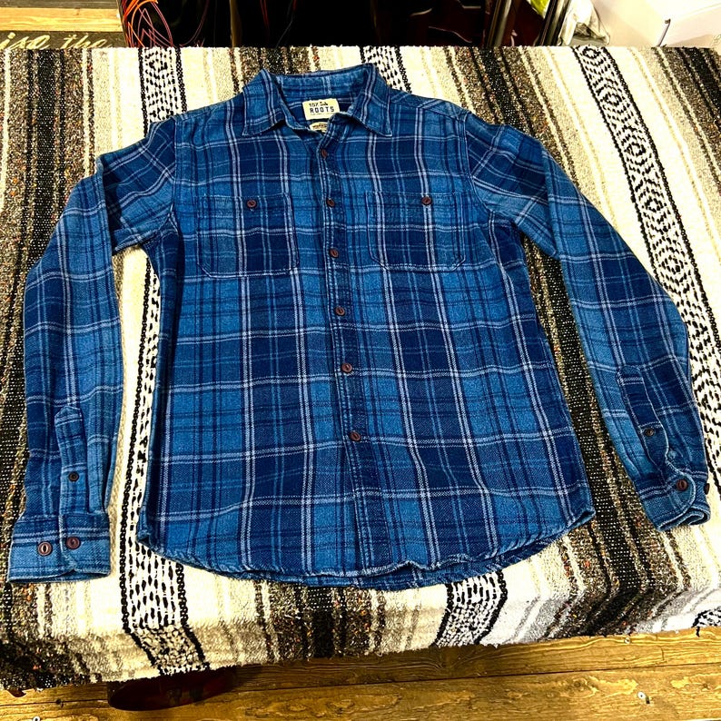May include: A blue plaid button-down shirt with long sleeves. The shirt features a collared neckline, two chest pockets, and buttoned cuffs. The shirt is made of a soft, flannel-like material. The brand name "Mesa Roots" is visible on the label.
