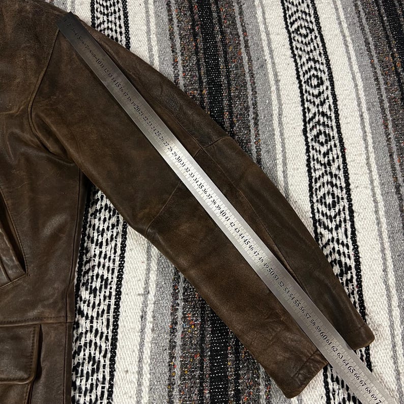 May include: A brown leather jacket with a ruler laid across the sleeve. The jacket has a worn appearance and features a pocket. The ruler measures up to 70 inches.