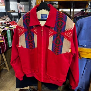 May include: A red zip-up jacket with a cream lining and a Pendleton label. The jacket has tan panels on the shoulders and chest, decorated with a geometric pattern in blue, green, and red. It features two front pockets and a ribbed hem.