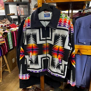 May include: A black zip-up jacket with a geometric pattern in grey, orange, purple, and green. The jacket has a black collar and cuffs, and a label is visible on the inside. The jacket is hanging on a wooden rack.