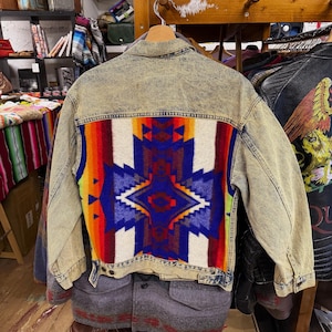 May include: A vintage denim jacket with a colourful, patterned textile panel on the back. The panel features a geometric design in shades of red, orange, blue, and white. The jacket has a faded, acid-wash appearance.