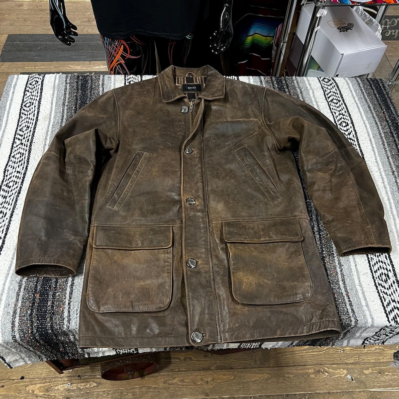 May include: A brown leather jacket with a button-up front, two chest pockets, and two lower pockets. The jacket has a collar and a zipper closure. The leather has a distressed appearance.