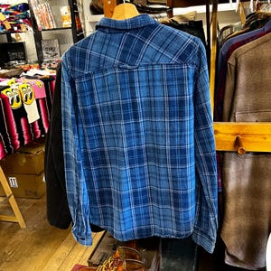 May include: A blue plaid button-down shirt with long sleeves. The shirt features a classic plaid pattern in shades of blue and white. The shirt is hanging on a wooden hanger, showcasing the back of the garment. The shirt is made of a soft, comfortable material.