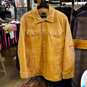 Könnte beinhalten: Hellbraune Lederjacke mit Knopfleiste, zwei Brusttaschen und zwei unteren Taschen. Die Jacke hat einen Kragen und der Markenname "TRAPPER" ist auf dem Etikett zu sehen. Die Jacke hängt an einem schwarzen Kleiderbügel.