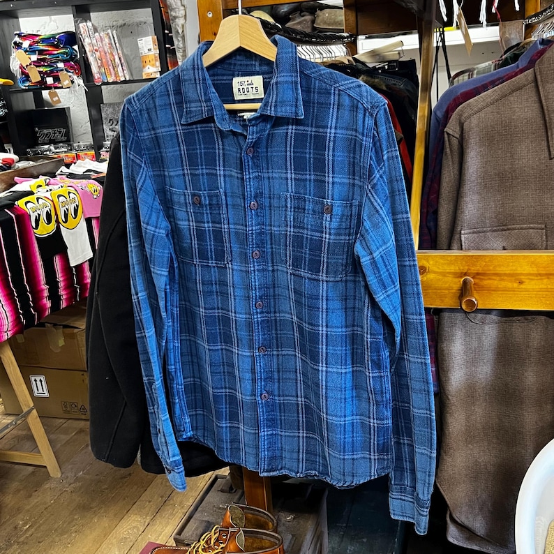 May include: A blue plaid button-down shirt with two chest pockets and the "Roots" logo on the label. The shirt is hanging on a wooden hanger. The shirt has a classic collar and buttoned cuffs.
