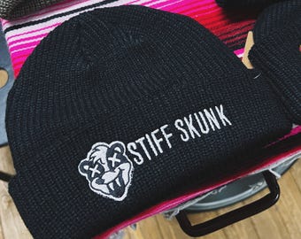 May include: Black knit beanie with the embroidered text "STIFF SKUNK" and a cartoon skunk graphic. The beanie is displayed on a striped pink and white fabric. Other beanies are visible in the background.