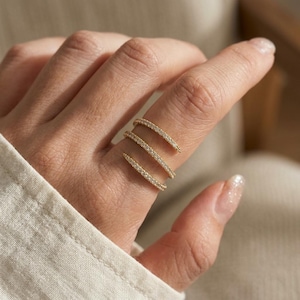 Op de afbeelding: Een goudkleurige spiraalring met drie afzonderlijke banden die om de vinger wikkelen. Elke band is ingelegd met een reeks kleine, heldere, sprankelende steentjes die het licht vangen en een continu glinsterend effect over de vinger creëren.