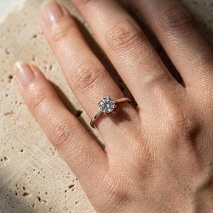 Könnte beinhalten: Ein Solitärring mit einem runden, im Brillantschliff geschliffenen, klaren Edelstein, der in eine polierte, silberfarbene Metallschiene gefasst ist. Der Ring wird am Finger getragen und zeigt die Krappenfassung, die den Stein über der Schiene hält.