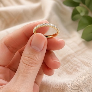 Può includere: Un anello a fascia color oro caratterizzato da una fila continua di piccole pietre chiare e scintillanti incastonate sulla superficie esterna. L'anello presenta una finitura metallica lucida e un profilo circolare sottile dal design uniforme.