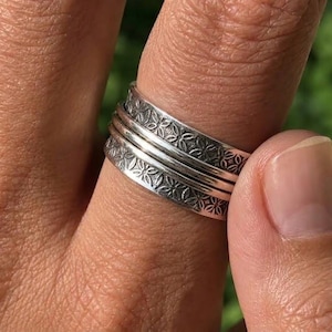 May include: A wide silver spinner ring featuring a repeating floral pattern engraved on the outer bands. The ring consists of a central smooth band flanked by two textured bands that rotate independently around the main body of the silver ring.