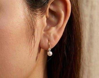 Könnte beinhalten: Ein goldener Ohrhänger mit einem kleinen, klaren, rund geschliffenen Edelstein, der über einer einzelnen, glatten, weißen, kugelförmigen Perle sitzt. Der Ohrring ist an einem dünnen Golddraht befestigt, der durch das Ohrläppchen geführt wird.