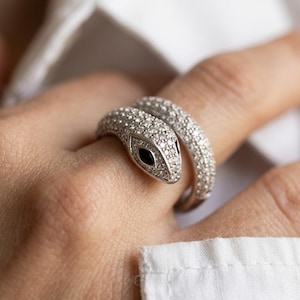 May include: A silver-toned snake ring featuring a pavé-set surface of small clear stones covering the entire band. The snake head is detailed with two marquise-cut black stones for eyes, creating a wrap-around design that encircles the finger.