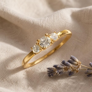 May include: A gold ring featuring three round-cut clear stones set in a row. The center stone is larger than the two flanking stones, and the band is a smooth, polished gold metal. The ring is positioned at an angle, showcasing the setting.