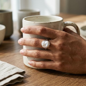 May include: A silver ring featuring a rectangular, textured white pearl set in a metal band. The ring is worn on a finger, with the hand resting against a speckled, off-white ceramic mug. The design highlights the organic shape of the pearl.