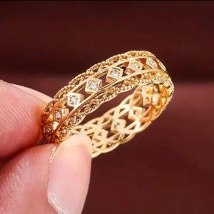 May include: A gold band ring featuring an intricate openwork lattice design with a series of small, clear, round-cut stones set within square-shaped metal frames. The edges of the band are finished with a delicate, beaded texture throughout.