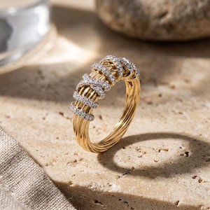 May include: A gold ring featuring a multi-strand cable design with a central section adorned with small, clear pavé-set stones. The band has a polished finish and a rounded profile, showcasing a textured, twisted metal appearance throughout the piece.
