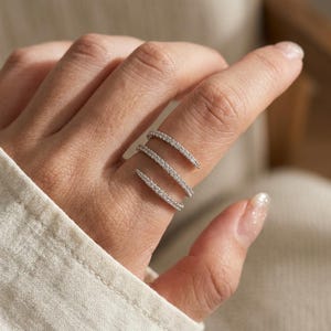 Puede incluir: Un anillo en espiral de tono plateado con tres bandas distintas que se envuelven alrededor del dedo. Cada banda está incrustada con pequeñas piedras claras y brillantes que captan la luz, creando una apariencia continua y superpuesta.
