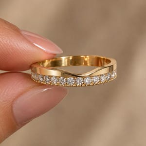 May include: A gold band ring featuring a curved, notched top edge and a row of small, round, clear, faceted stones embedded along the lower half of the band, creating a sparkling, channel-set appearance with a polished, reflective metal finish.