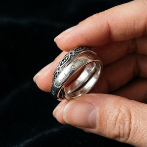 Può includere: Un anello in argento composto da due fasce intrecciate. Una fascia presenta una superficie liscia e lucida con la parola LOVE incisa in lettere maiuscole. La seconda fascia mostra un motivo complesso, scuro e strutturato con piccoli dettagli circolari.