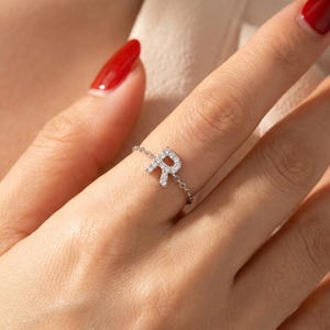 Peut inclure: Une bague chaîne argentée ornée d'un pendentif en forme de lettre R majuscule, incrusté de petites pierres claires et scintillantes. La lettre R est placée au centre de l'anneau en fine chaîne métallique qui entoure le doigt avec éclat.