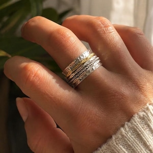 May include: A multi-band spinner ring featuring a combination of silver and gold-toned metal. The design includes a central twisted rope texture, flanked by two wider bands with a hammered, dimpled finish, creating a layered appearance.