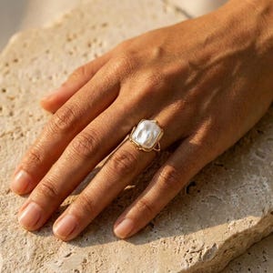May include: A gold ring featuring a single, irregular, white baroque pearl set within a delicate, thin gold wire frame that wraps around the pearl, securing it in place on the finger with a minimalist and open-ended metal band design.