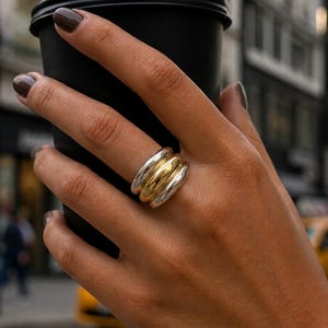Peut inclure: Une bague multi-anneaux composée de trois bandes métalliques épaisses, arrondies et polies. La bande centrale est dorée, tandis que les deux bandes extérieures sont argentées. La bague présente une surface bombée et lisse.