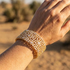 May include: A wide gold-tone cuff bracelet featuring an intricate lattice design encrusted with numerous clear, sparkling stones in various shapes, including round, pear, and rectangular cuts, creating a dense, textured, and reflective surface.