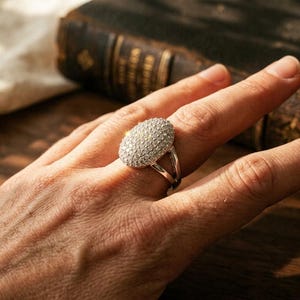 May include: A silver-toned ring featuring a large, oval-shaped setting densely encrusted with numerous small, clear, sparkling stones. The band of the ring is split into two distinct sections that meet at the base of the oval setting.