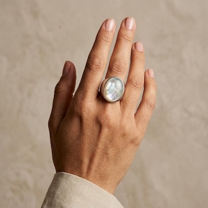 Può includere: Un anello in argento con una grande pietra cabochon ovale in madreperla iridescente, liscia e lucida, montata su una fascia in metallo. L'anello è indossato sul dito medio, mettendo in risalto la superficie perlacea e riflettente.