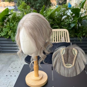 May include: A light ash blonde hair topper styled with soft waves on a mannequin head. The underside features a brown mesh base with three metal pressure-sensitive clips attached to the perimeter for secure placement on the scalp.