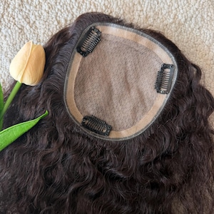 May include: A dark brown hair topper featuring a circular beige mesh base with three black metal pressure-sensitive clips attached to the perimeter. The hair has a wavy texture and is positioned next to a single pale yellow tulip flower.