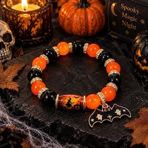 Könnte beinhalten: Ein elastisches Armband mit abwechselnd orangefarbenen und schwarzen Perlen, goldenen Strass-Abstandshaltern und einer zentralen zylindrischen Perle mit Hexen- und Kürbis-Motiv. Ein schwarzer fledermausförmiger Anhänger mit zwei Geistern und roten Akzenten.
