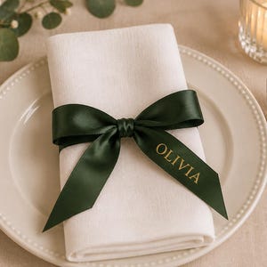 May include: A white fabric napkin folded on a cream-coloured plate, tied with a dark green satin ribbon. The ribbon features the name OLIVIA printed in gold lettering on one of the tails. The setting includes a silver fork and a silver knife.