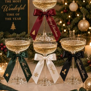 May include: Four clear, ribbed coupe glasses filled with a sparkling beverage. Each glass features a satin ribbon tied around the stem. The ribbons are burgundy, dark green, white, and black, printed with gold text and icons including Merry Christmas, The Smiths, Sophie, and Christmas Cheers.
