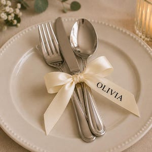 May include: A stainless steel fork, knife, and spoon set resting on a cream-coloured ceramic plate with a beaded rim. The cutlery is tied together with a cream satin ribbon featuring the name OLIVIA printed in black capital letters.