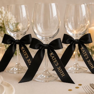 May include: Three clear glass wine glasses, each featuring a black satin ribbon tied around the stem. The ribbons are printed with the gold text KATE'S 30TH. The glasses have a standard bowl shape, a slender stem, and a circular base for stability.