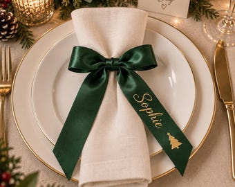 May include: A beige linen napkin folded and secured with a dark green satin ribbon tied into a bow. The ribbon features the name Sophie printed in gold script lettering, accompanied by a small gold Christmas tree icon, centred on a white plate.