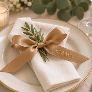 May include: A white ceramic plate with a gold rim features a folded white cloth napkin topped with a sprig of green leaves and a gold satin ribbon tied in a bow. The ribbon is printed with the name EMMA in gold capital letters.