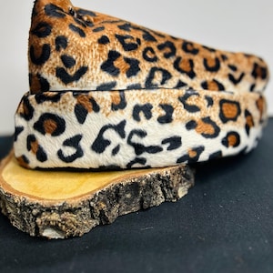Puede incluir: Una diadema con un patrón de estampado de leopardo texturizado con manchas negras, marrones y color crema. El material tiene una apariencia corta similar al pelaje y está formado por una banda ancha y redondeada que se curva hacia arriba.