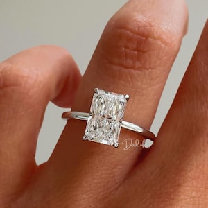 May include: A silver-toned ring featuring a large, rectangular radiant-cut clear gemstone set in a four-prong setting on a thin, polished metal band. The centre stone displays multiple facets that reflect light, creating a brilliant appearance.