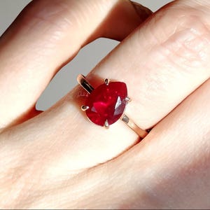 Puede incluir: Un anillo con una piedra preciosa de color rojo intenso en forma de pera, montada en una banda de metal de tono dorado pulido. La piedra está asegurada por cuatro pequeñas garras y muestra una superficie facetada que refleja la luz.