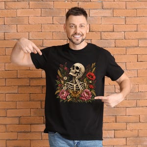 May include: A black t-shirt featuring a graphic print of a human skull and upper torso skeleton surrounded by colorful flowers, including red poppies and pink peonies, with a moth positioned at the base of the floral arrangement.