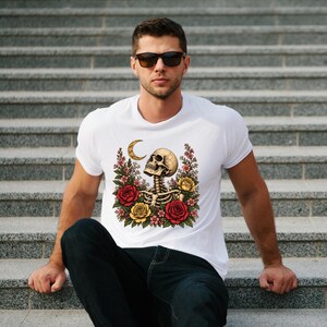 May include: A white short-sleeved t-shirt featuring a graphic print of a human skull and ribcage surrounded by red and yellow roses and a crescent moon. The design is centered on the chest of the garment, which is worn with dark blue jeans.