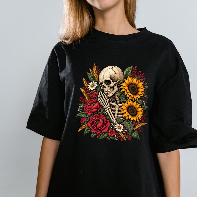 May include: A black t-shirt featuring a graphic print of a human skull surrounded by a vibrant floral arrangement. The design includes bright yellow sunflowers, deep red peonies, small white daisies, green leaves, and stalks of golden wheat.