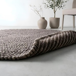 May include: A round, taupe-colored rug constructed from numerous small, densely packed felted wool balls. The rug is partially folded over, revealing a structured, grid-like backing material underneath the textured, spherical surface.