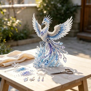 May include: A beaded phoenix sculpture featuring white, blue, and purple crystals, perched on a translucent blue crystal base. The set includes loose beads, metal scissors, wire, and small snowflake-shaped charms arranged on a wooden surface.