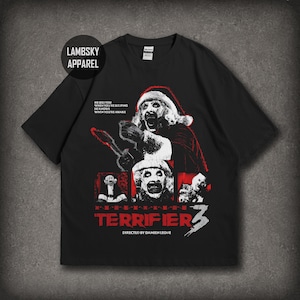 Può includere: Una t-shirt nera a maniche corte con una stampa grafica di Art the Clown con un cappello da Babbo Natale. Il design include il testo HE SEES YOU WHEN YOU'RE SLEEPING HE KNOWS WHEN YOU'RE AWAKE, TERRIFIER 3 e DIRECTED BY DAMIEN LEONE in rosso e bianco.