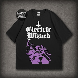 May include: A black short-sleeve t-shirt featuring the white text Electric Wizard with an inverted cross symbol above it. Below the text is a purple graphic illustration of a figure with a skull and skeletal elements on a black background.